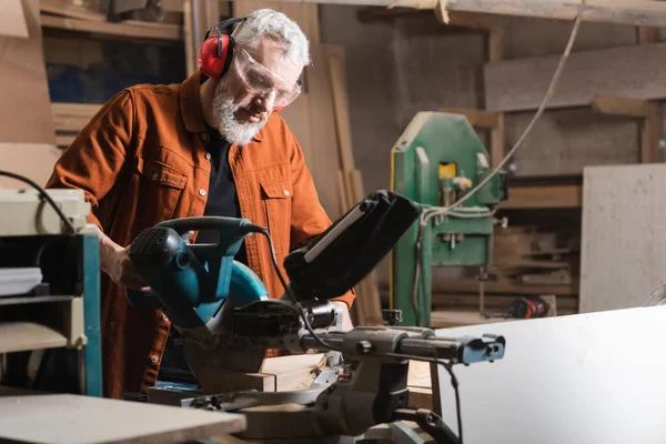 Menuisier barbu en lunettes de travail sur scie à onglets en atelier — Photo de stock