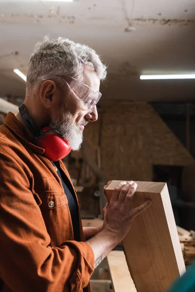 Positive furniture designer in goggles checking wooden plank in workshop — Stock Photo