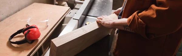 Partial view of woodworker with board near safety earmuffs in workshop, banner — Stock Photo