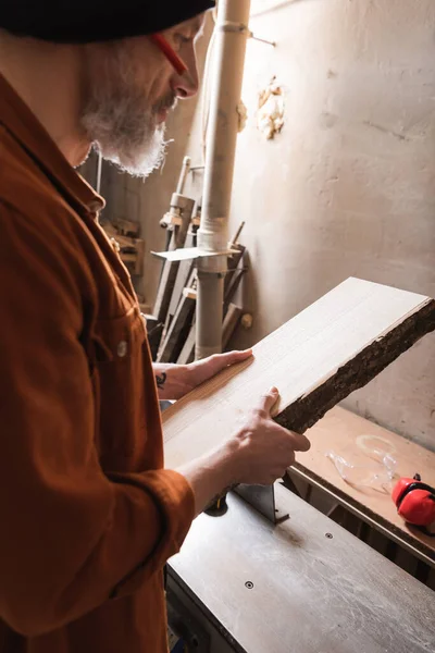 Bearded woodworker in beanie holding board in workshop — Stock Photo