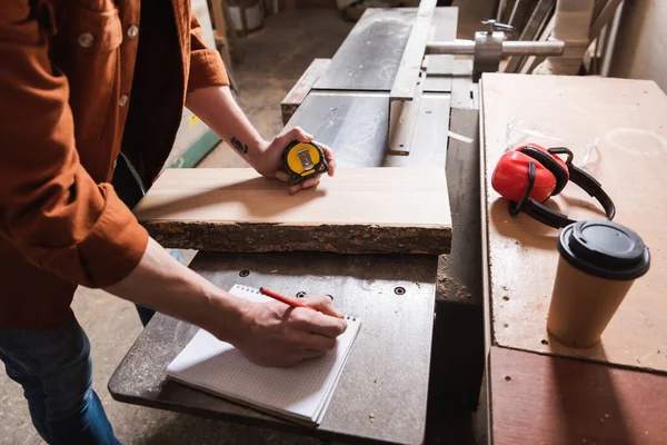 Cropped view of carpenter writing in notebook near coffee to go on workbench — Stock Photo