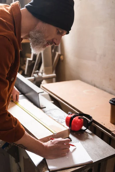 Bearded carpenter in beanie measuring board and writing in notebook — Stock Photo