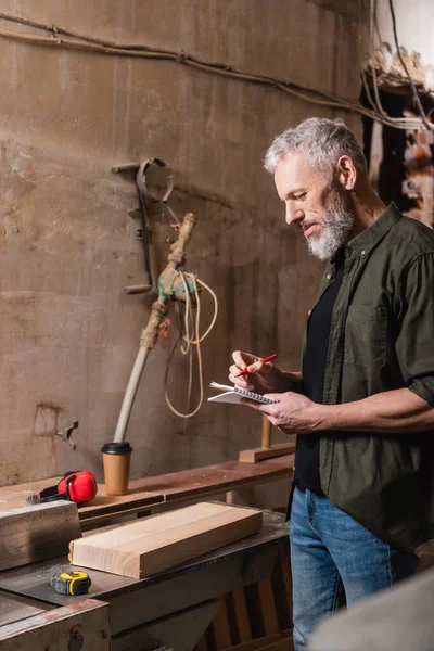 Side view o bearded carpenter with pencil and notebook near board on workbench — Stock Photo