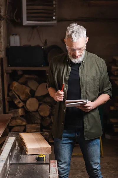 Designer de meubles barbu en lunettes debout avec cahier et crayon dans l'atelier — Photo de stock