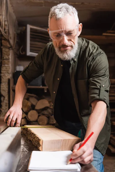 Mature furniture designer measuring board and writing in notebook — Stock Photo