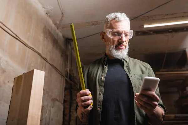 Bearded carpenter in goggles holding tape measure and smartphone in workshop — Stock Photo