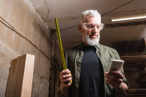 Carpinteiro barbudo em óculos segurando fita métrica e smartphone na oficina — Fotografia de Stock