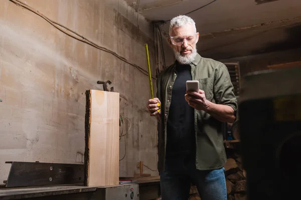 Бородатый дизайнер мебели, стоящий с магнитофоном и смартфоном в столярной мастерской — стоковое фото