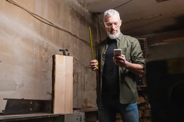 Bearded furniture designer standing with tape meter and smartphone in carpentry studio — Stock Photo