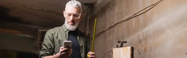 Menuisier barbu avec ruban à mesurer en utilisant smartphone en atelier, bannière — Photo de stock