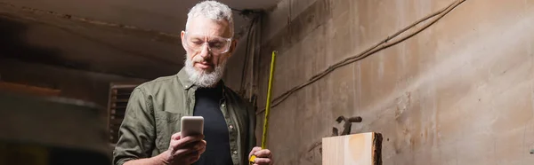 Menuisier barbu avec ruban à mesurer en utilisant smartphone en atelier, bannière — Photo de stock