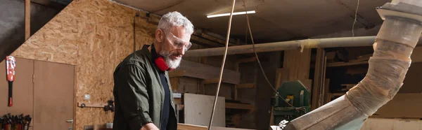 Middle aged carpenter in goggles working in woodwork studio, banner — Stock Photo