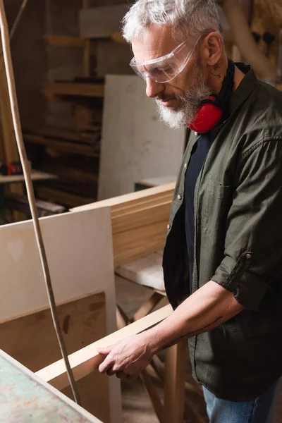 Зрелый плотник в очках держит деревянную доску в мастерской по деревообработке — стоковое фото