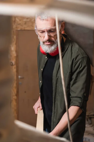 Bearded furniture designer in goggles working in woodwork studio on blurred foreground — Stock Photo