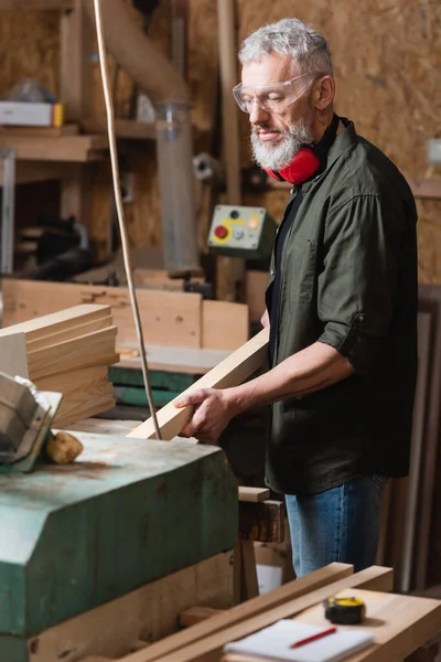 Menuisier barbu tenant une planche de bois tout en travaillant dans un atelier de menuiserie — Photo de stock