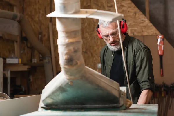 Menuisier en lunettes et cache-oreilles travaillant près d'une hotte de ventilation floue — Photo de stock