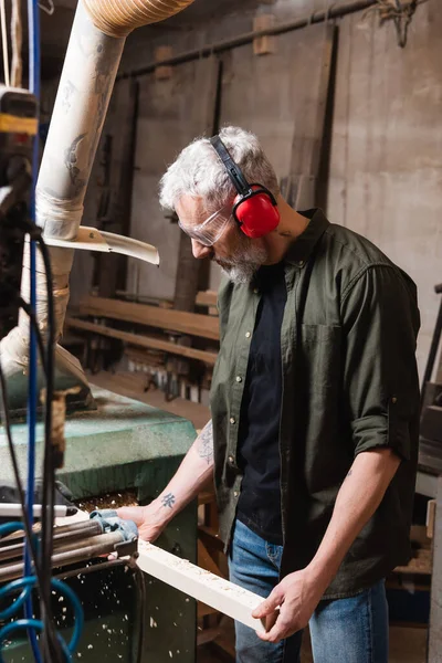 Carpintero maduro en gafas de trabajo con tablón de madera en taller - foto de stock