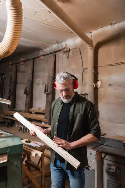 Carpinteiro em óculos e auriculares verificando prancha de madeira na oficina — Fotografia de Stock