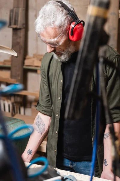 Tätowierte Möbeldesignerin mit Brille und schützenden Ohrenschützern bei der Arbeit im Tischleratelier — Stockfoto