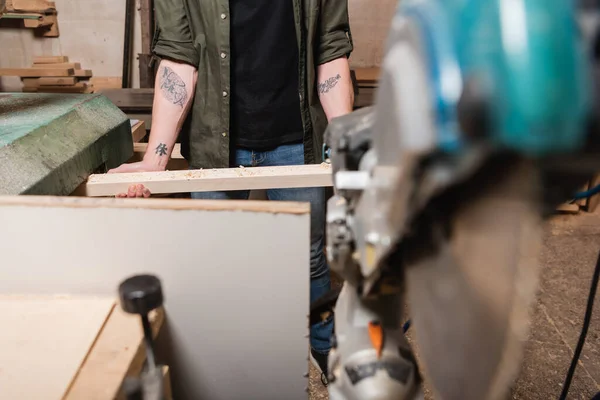 Частковий вид татуйованого працівника деревини, що тримає дошку під час роботи на розмитому передньому плані — стокове фото