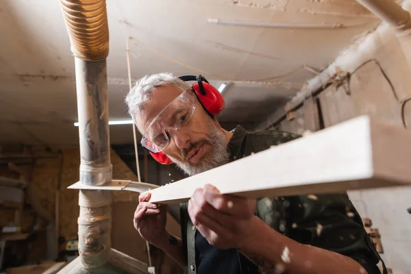 Marceneiro em googles e protetores auriculares verificando prancha na oficina — Fotografia de Stock