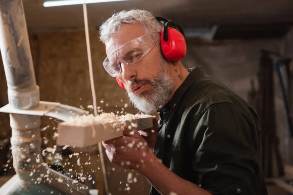 Carpenter in goggles and protective earmuffs blowing sawdust out of plank — Stock Photo