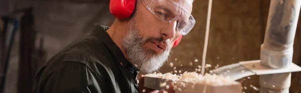 Falegname maturo in occhiali che soffia segatura da tavola di legno, banner — Foto stock