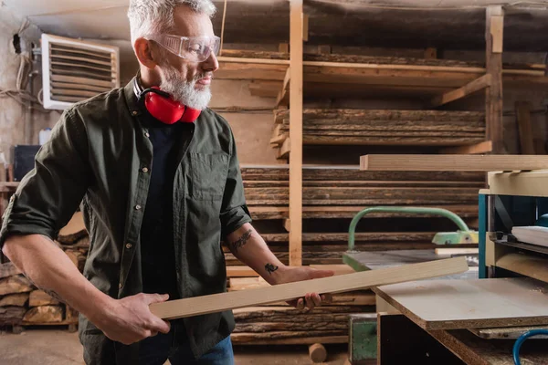 Carpenter in goggles holding board near thickness planer — Stock Photo