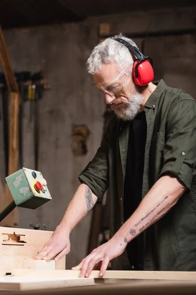 Charpentier barbu dans des cache-oreilles protecteurs travaillant sur une machine de menuiserie — Photo de stock