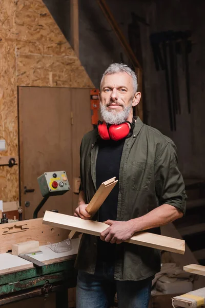 Bearded carpenter holding wooden details and looking at camera in workshop — Stock Photo