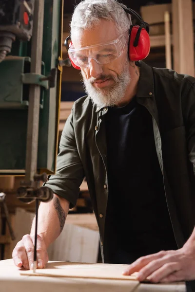 Menuisier barbu dans des lunettes de coupe de bois avec scie à ruban — Photo de stock