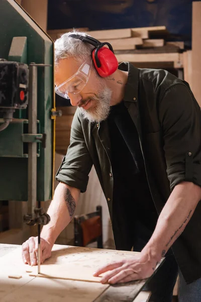 Bearded carpenter in goggles cutting plywood with band saw — Stock Photo