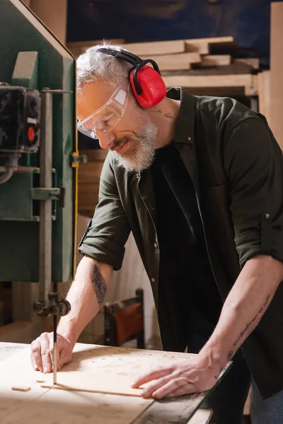 Menuisier barbu dans des lunettes de coupe contreplaqué avec scie à ruban — Photo de stock