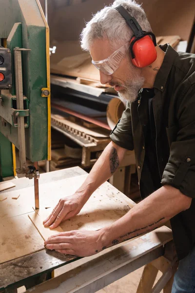 Tattooed carpenter in goggles cutting plywood with band saw — Stock Photo