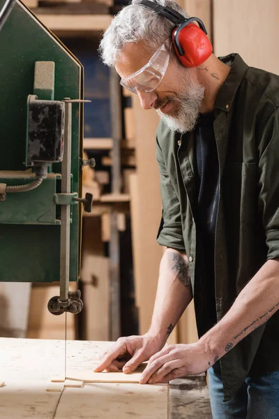 Positive carpenter in goggles cutting plywood on band saw — Stock Photo
