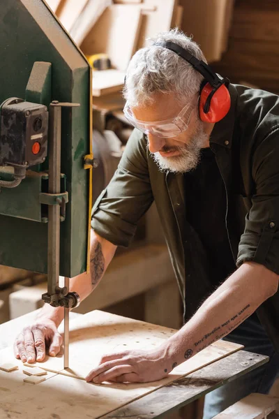 Bearded woodworker in goggles cutting plank on band saw — Stock Photo