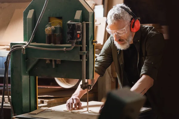 Grey haired carpenter cutting plywood on band saw in workshop — Stock Photo