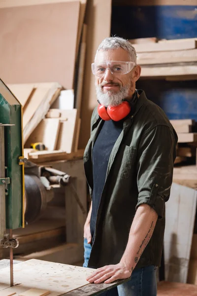 Mature menuisier souriant à la caméra près de scie à ruban dans atelier de menuiserie — Photo de stock