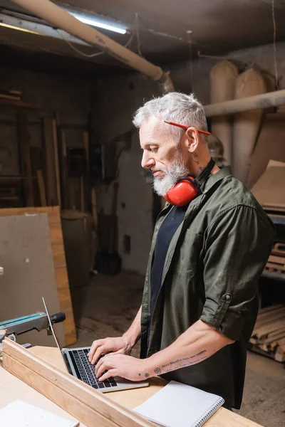 Side view of furniture designer typing on laptop in woodwork studio — Stock Photo