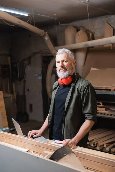 Designer de meubles barbu regardant la caméra près d'un ordinateur portable dans un studio de menuiserie — Photo de stock