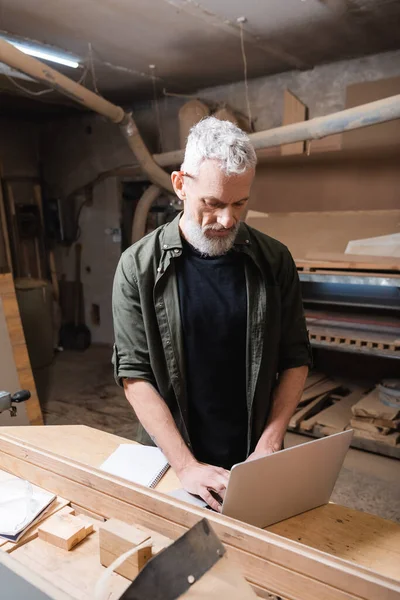 Bearded furniture designer typing on laptop in woodwork studio — Stock Photo