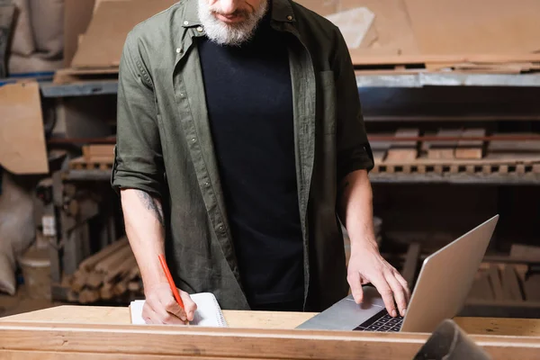 Cropped view of furniture designer writing in notebook near laptop on workbench — Stock Photo