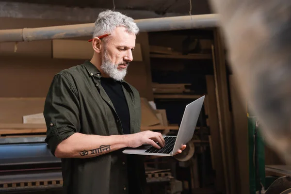 Designer de móveis de cabelos grisalhos em estúdio de madeira digitando no laptop em primeiro plano desfocado — Fotografia de Stock