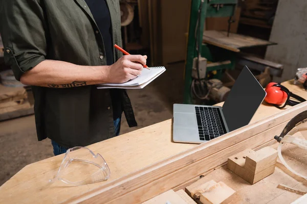 Cropped view of furniture designer writing in notebook near laptop with blank screen — Stock Photo