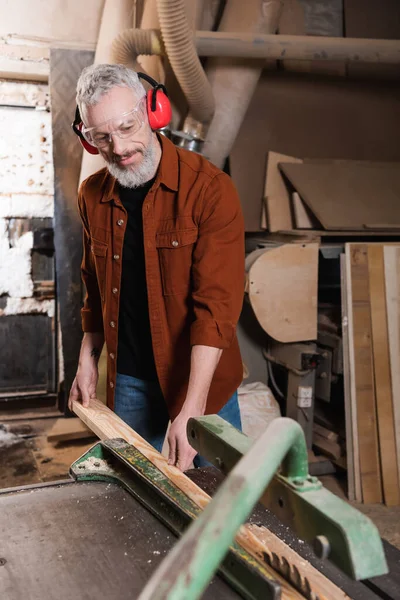 Carpenter in goggles and protective earmuffs holding plank near workbench — Stock Photo