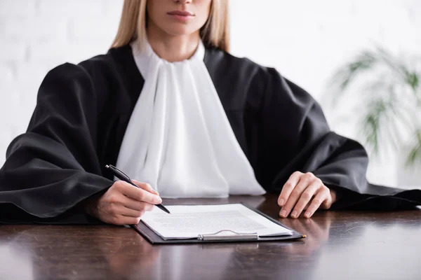 Vista cortada do promotor no manto sentado com caneta perto da área de transferência com ação judicial — Fotografia de Stock