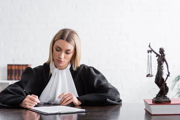 Prosecutor in black mantle signing lawsuit near blurred themis statue - foto de stock