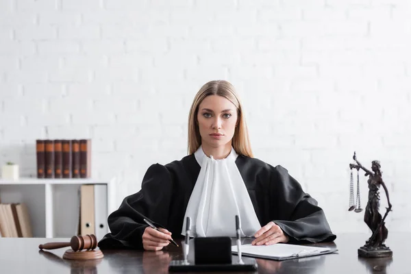 Judge in mantle looking at camera while sitting near gavel and themis statue — Fotografia de Stock