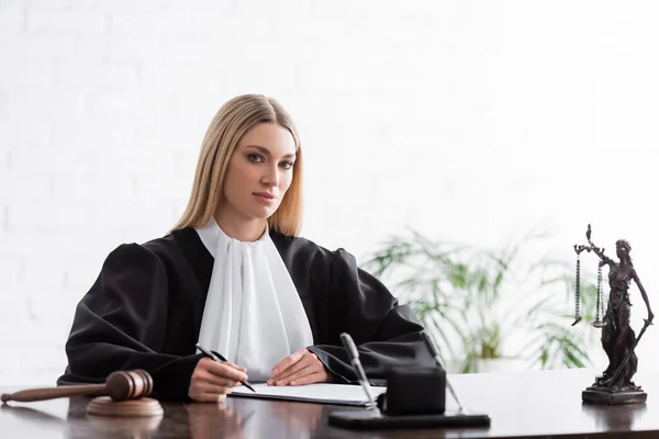 Blonde judge looking at camera while sitting with pen near gavel and blurred themis statue — Foto stock