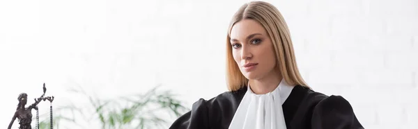 Blonde judge looking at camera near blurred themis statue, banner — Fotografia de Stock